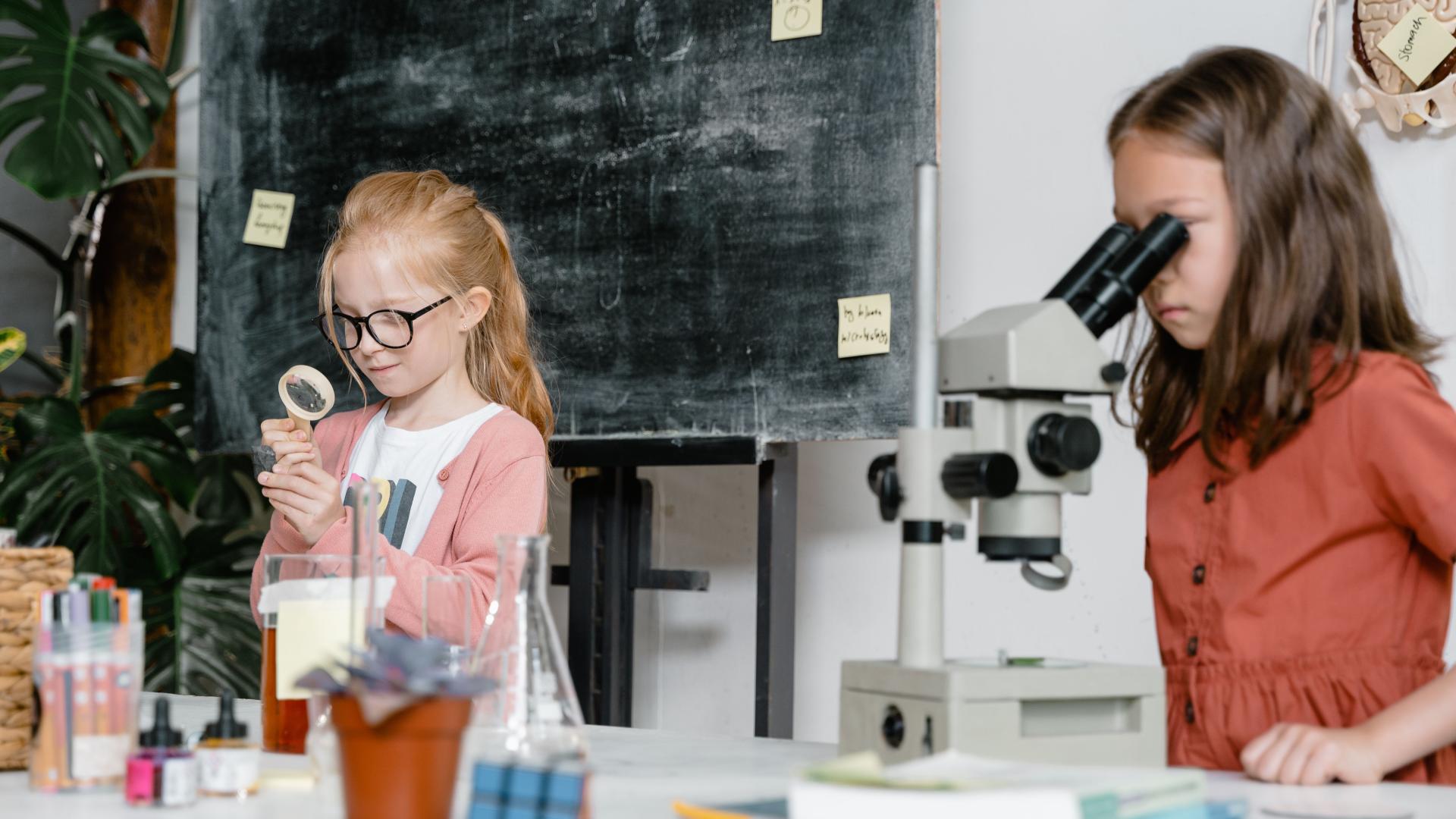 One elementary aged girl looking through a microscope while a second elementary ages girl looks closely at a plant with a handheld microscope