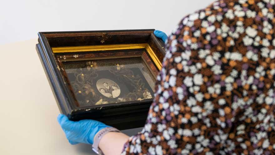 Archivist wearing blue latex gloves holds an old black and white portrait in an ornate frame