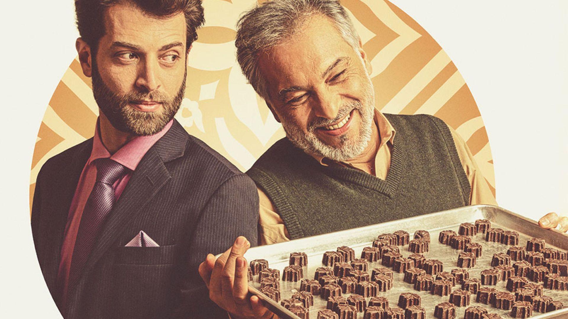 Two men standing side by side, one smiling and holding a tray of chocolates.
